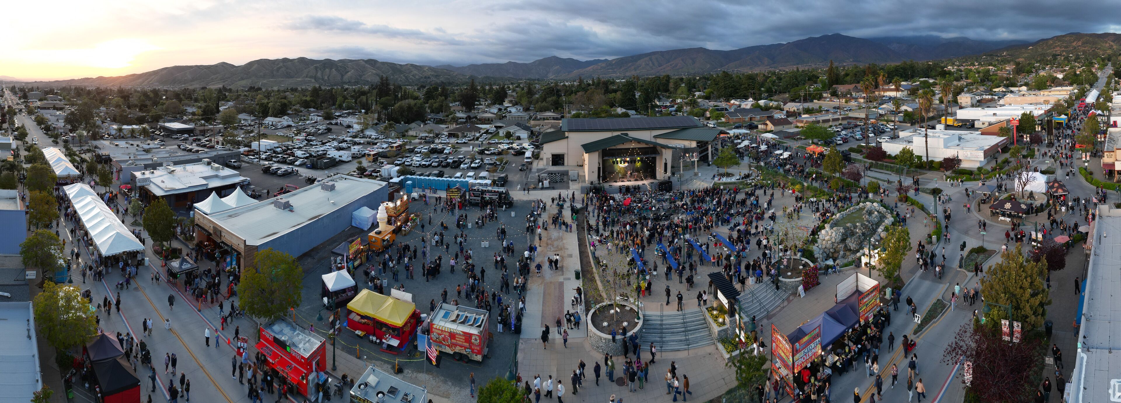 A bustling outdoor event with a stage, food stalls, and a crowd in a town square surrounded by mountains.
