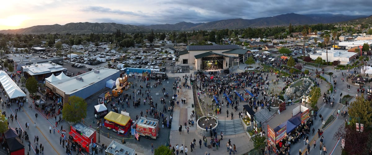 A bustling outdoor event with a stage, food stalls, and a crowd in a town square surrounded by mountains.