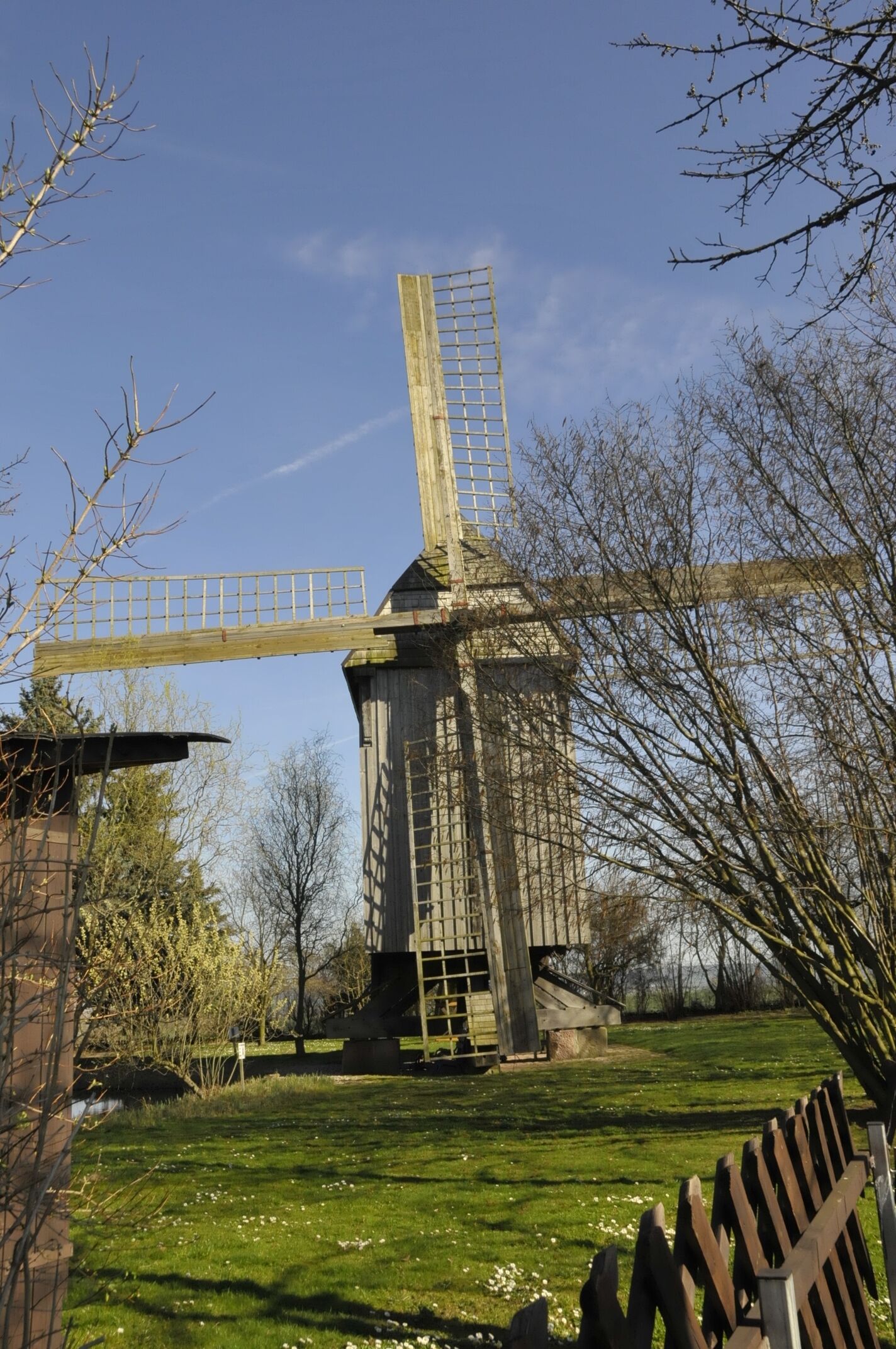 A windmill in a grassy field with trees and a wooden fence.