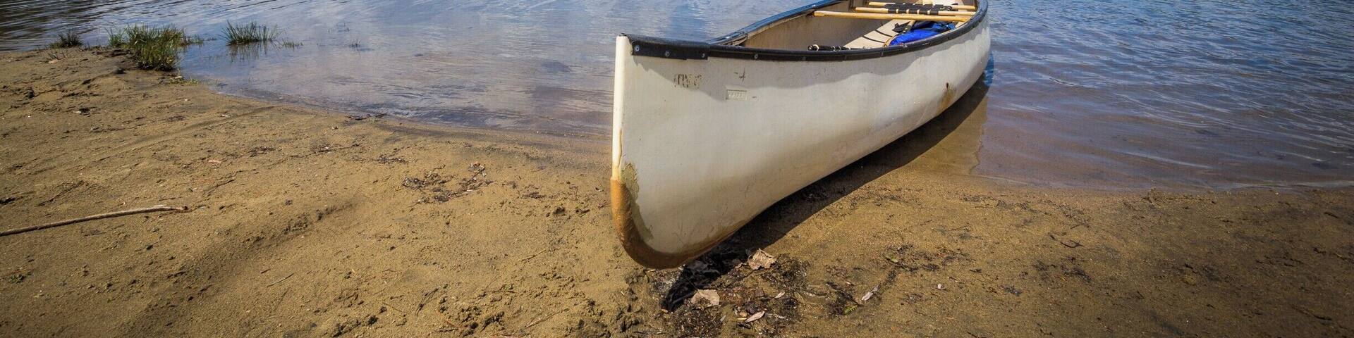A canoe on a sandy shore by a calm lake with trees in the background.