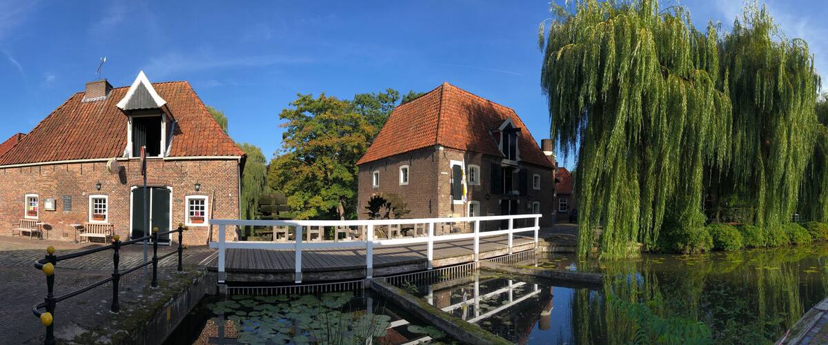A canal with a wooden bridge, a weeping willow tree, and brick buildings.