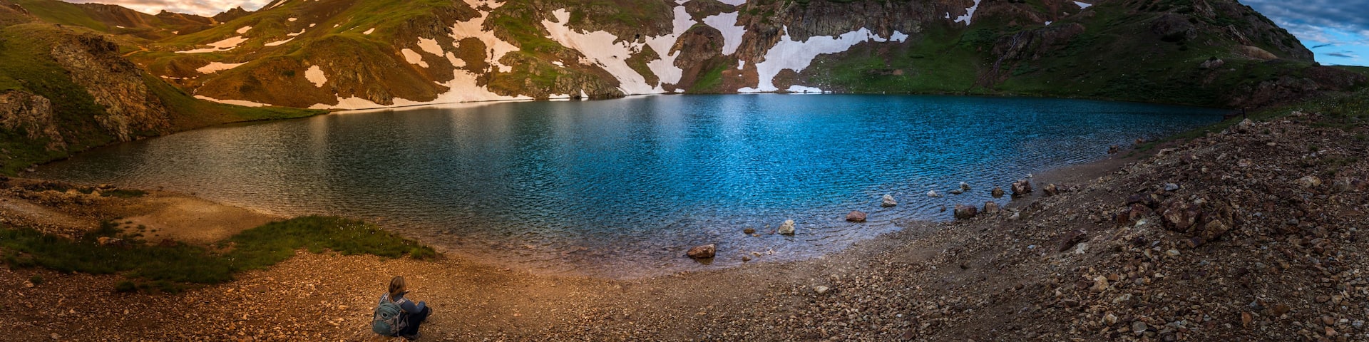 A mountain lake with snow patches, a rocky shoreline, and a cloudy sky.