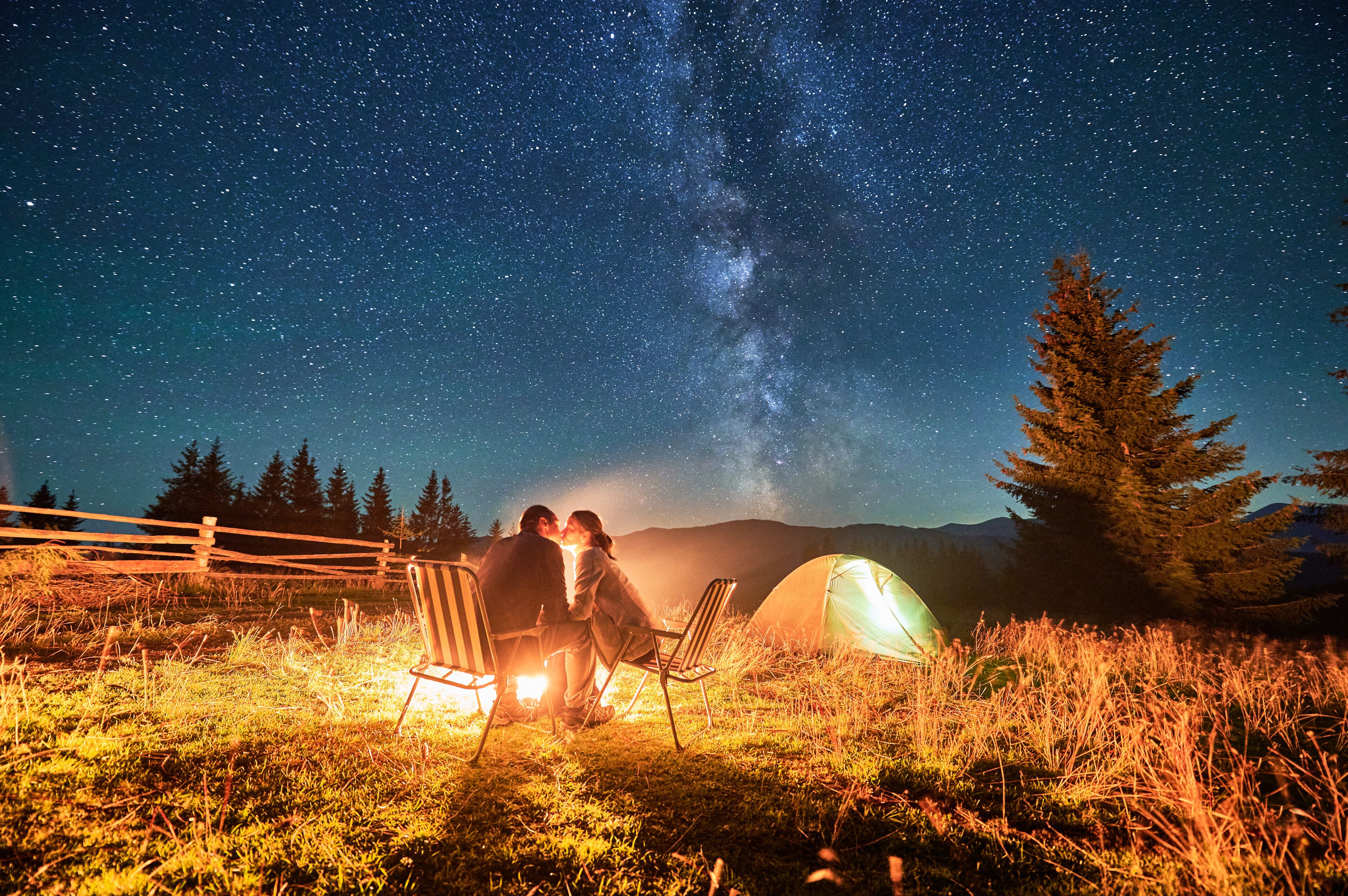 A couple under a starry sky with a Milky Way view, camping chairs, a tent, and a wooden fence in a grassy field.