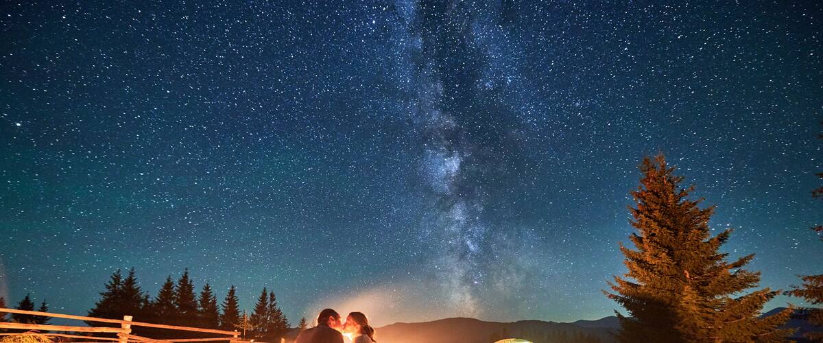 A couple under a starry sky with a Milky Way view, camping chairs, a tent, and a wooden fence in a grassy field.