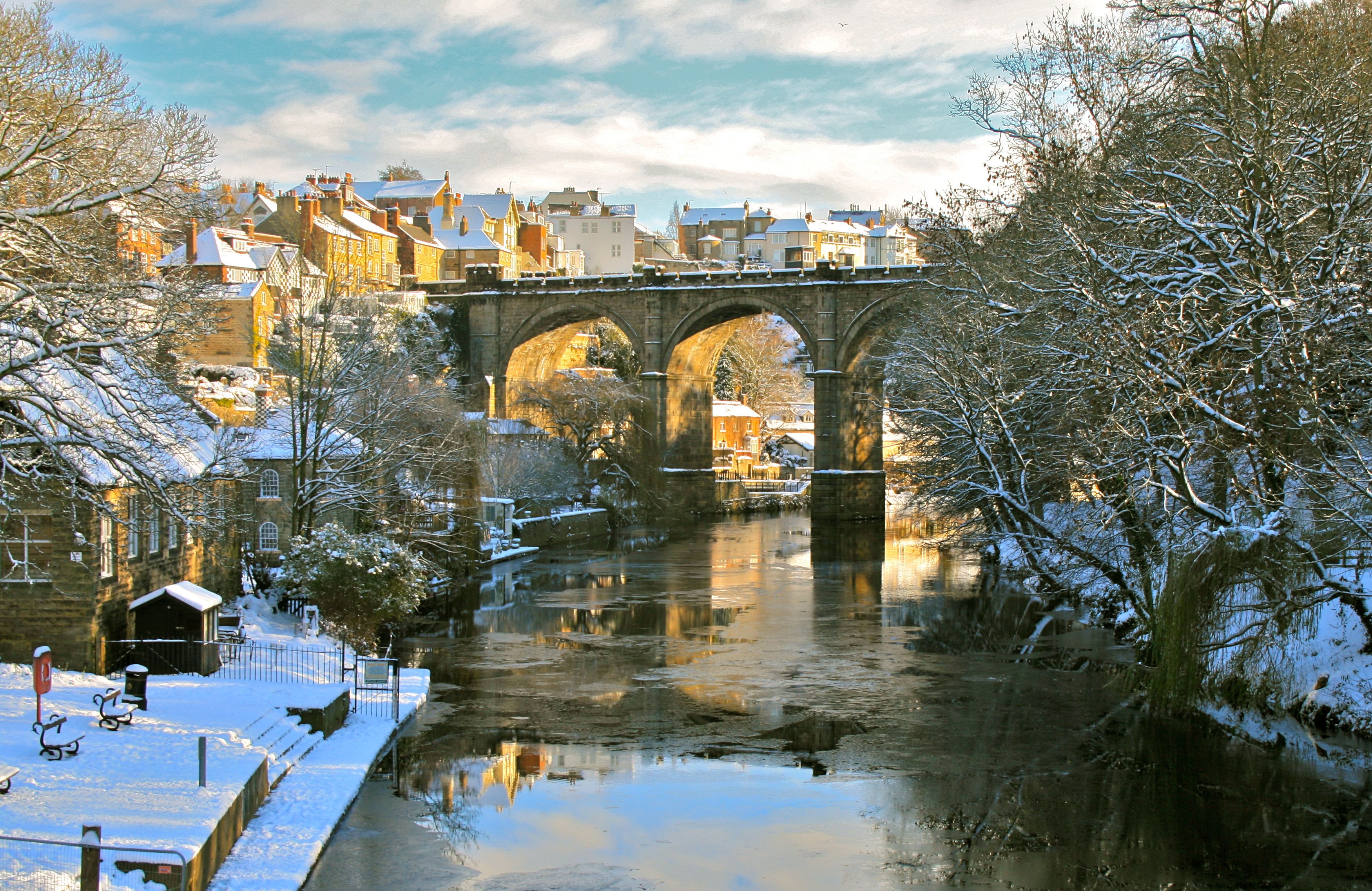 Un río cubierto de nieve con un puente de piedra, edificios y árboles sin hojas.