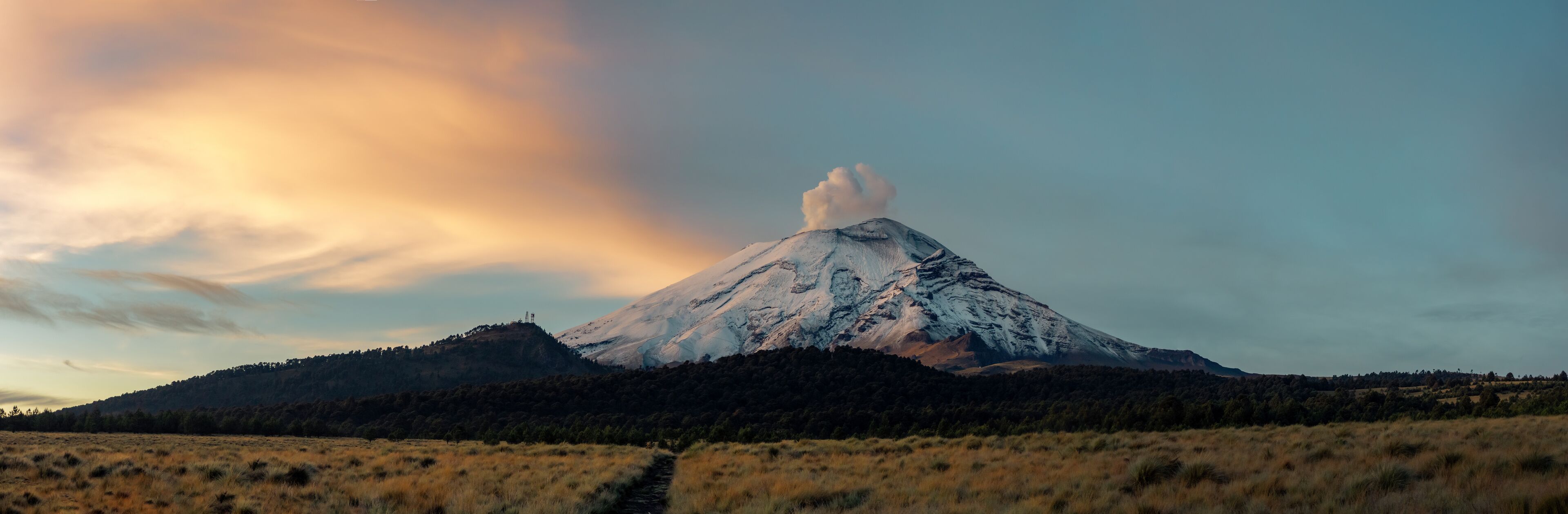 A snow-capped mountain with a visible smoke plume, surrounded by forested hills and a clear sky.