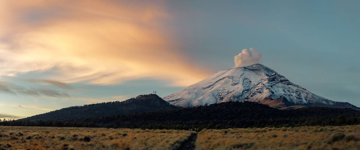 A snow-capped mountain with a visible smoke plume, surrounded by forested hills and a clear sky.