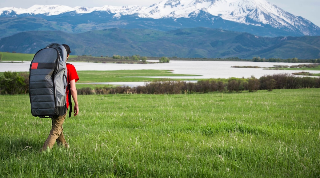 Young male hikes through grassy field towards a lake with an inflatable paddleboard in the mountains