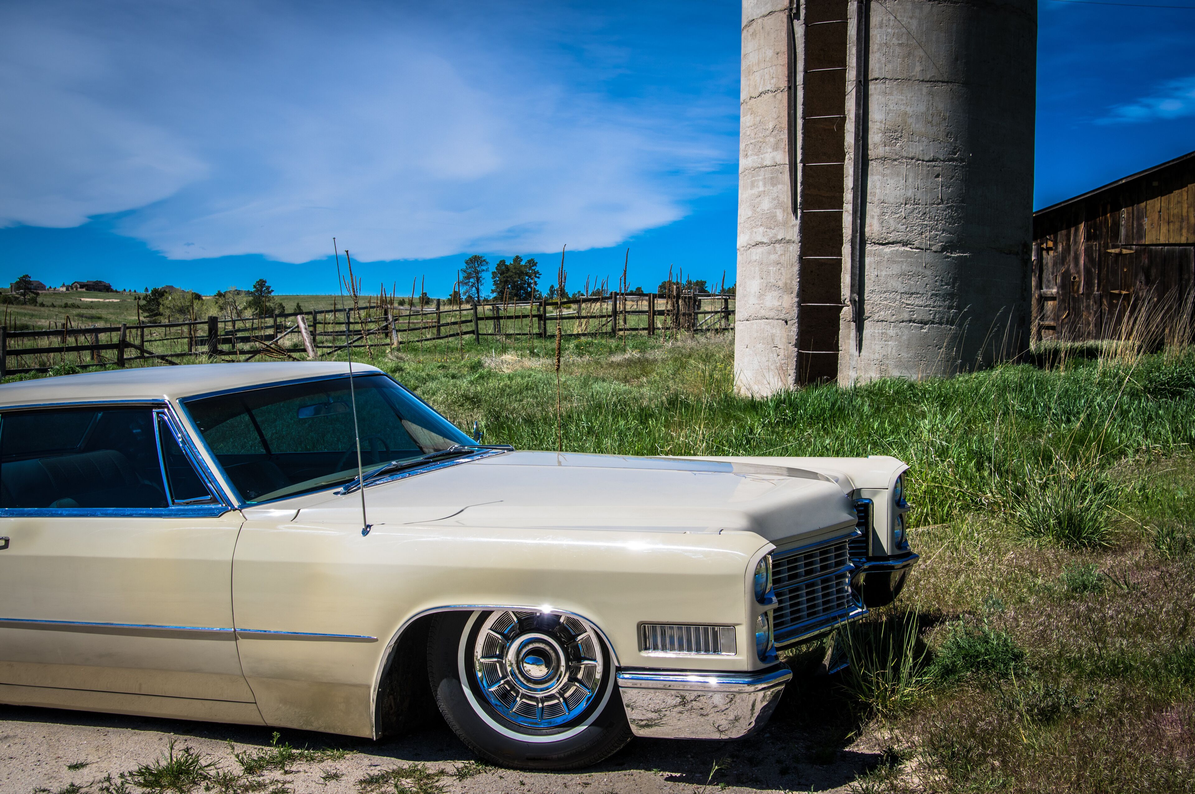 A vintage beige car parked near a concrete structure with a wooden fence and grassy field in the background.