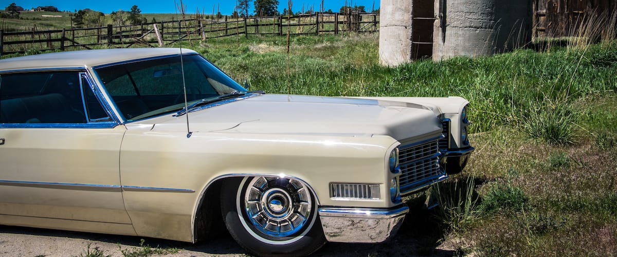 A vintage beige car parked near a concrete structure with a wooden fence and grassy field in the background.