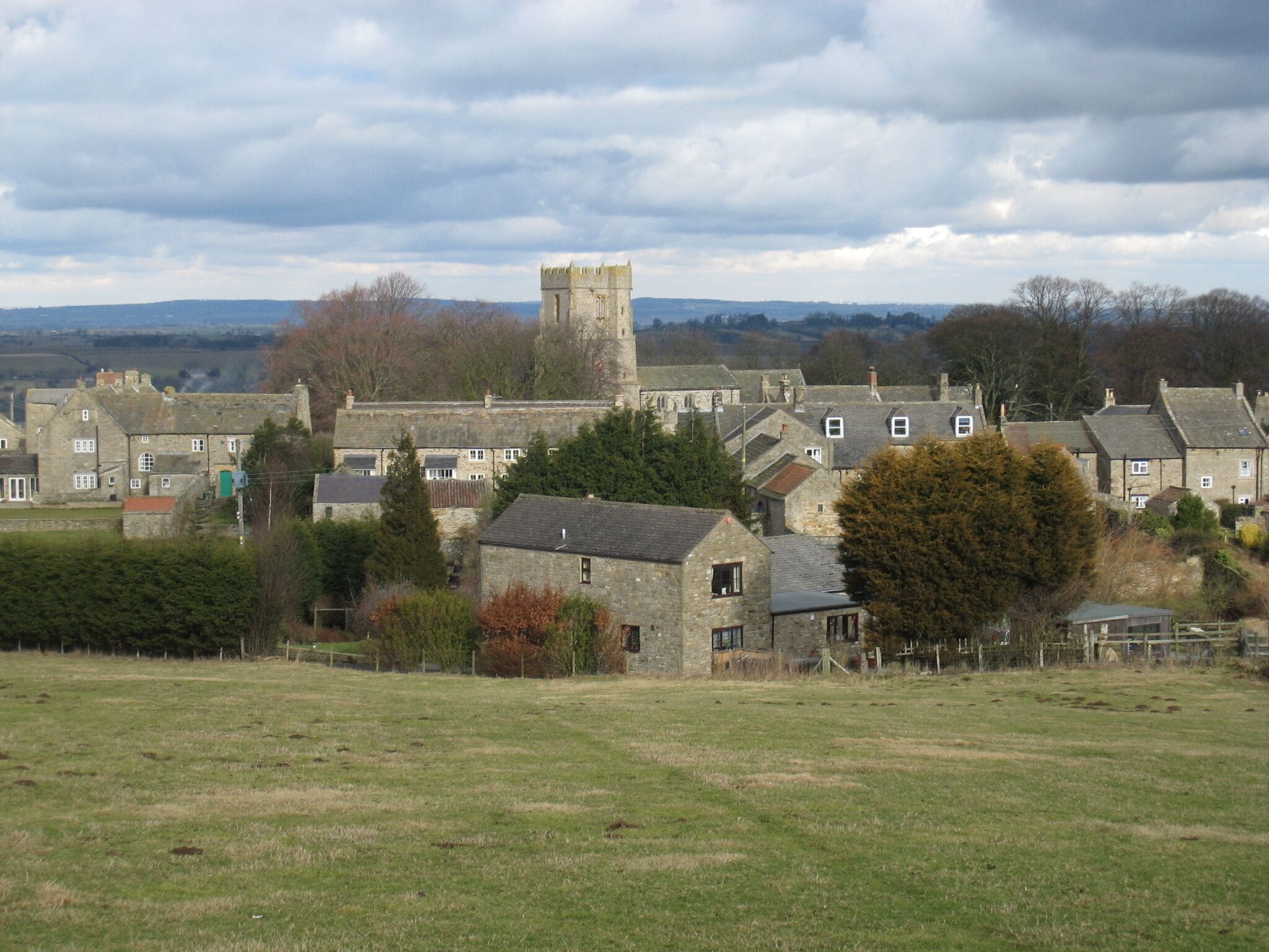 A historic stone building with a tower, surrounded by traditional houses and a grassy field.