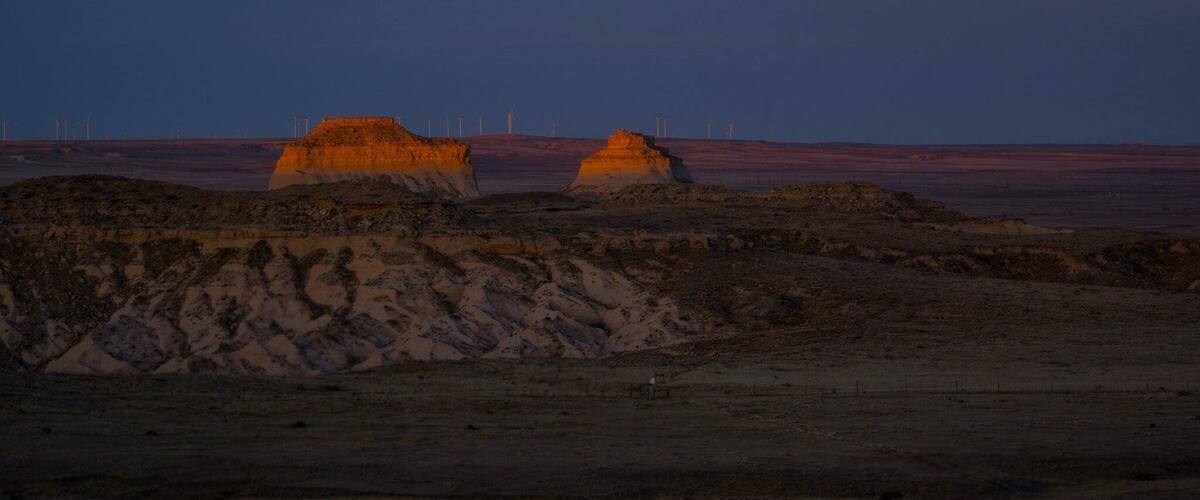 A desert landscape with two prominent rock formations and a series of wind turbines in the background.