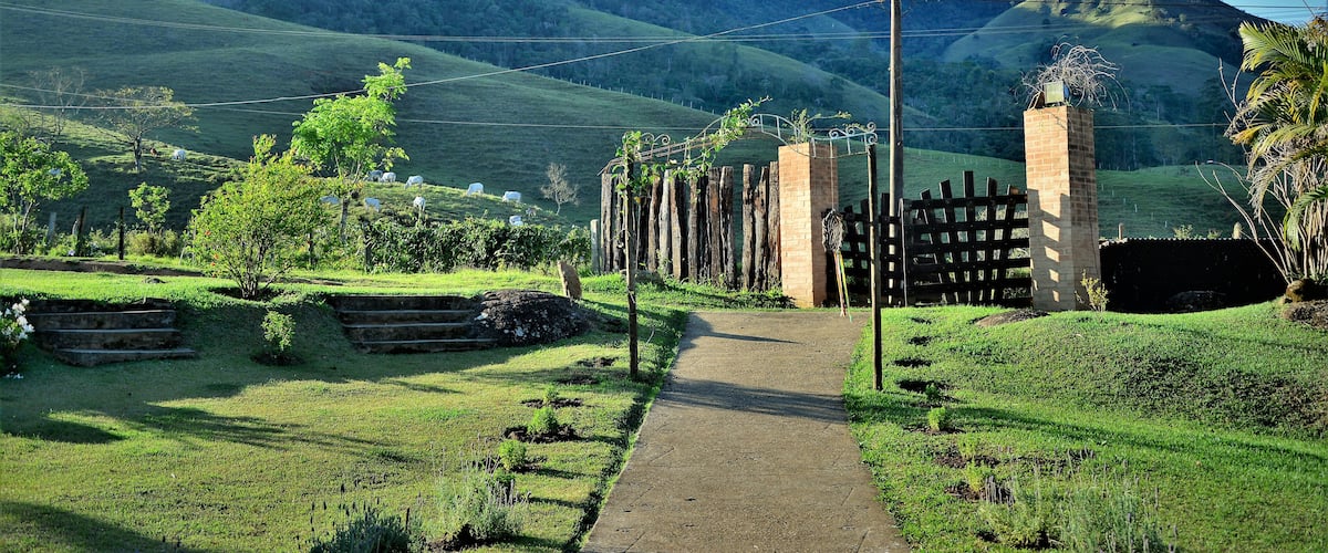 A rural landscape with a dirt path leading to a gated entrance, surrounded by lush green fields and hills in the background.