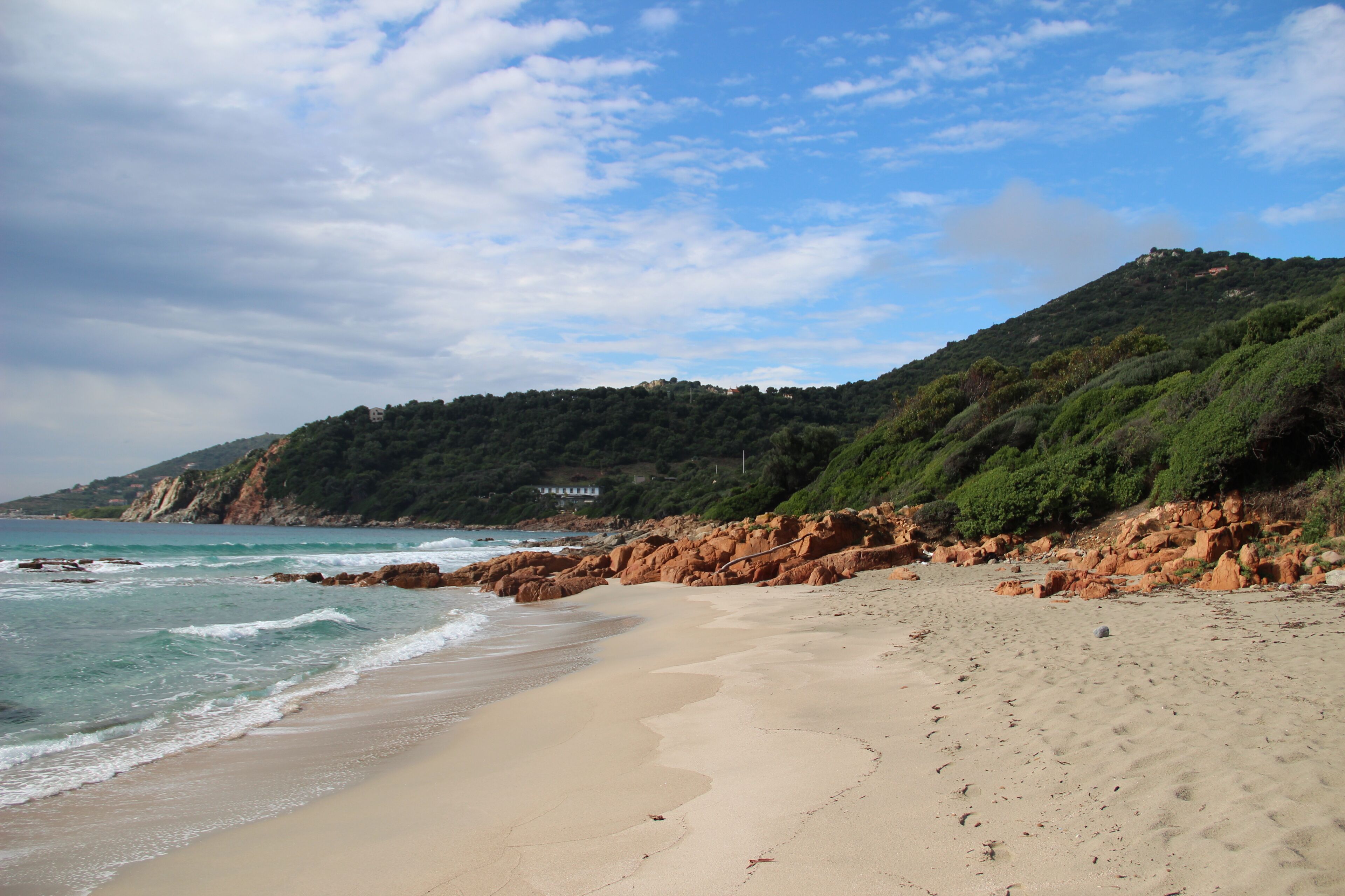 A sandy beach with gentle waves, a rocky coastline, and a hill with dense vegetation.