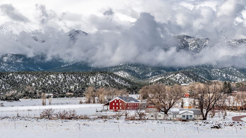 A snow-covered landscape with a red barn, a white house, and snow-capped mountains in the background.