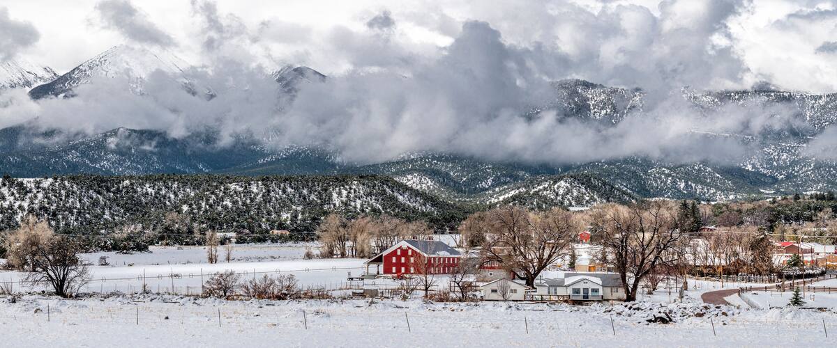 A snow-covered landscape with a red barn, a white house, and snow-capped mountains in the background.