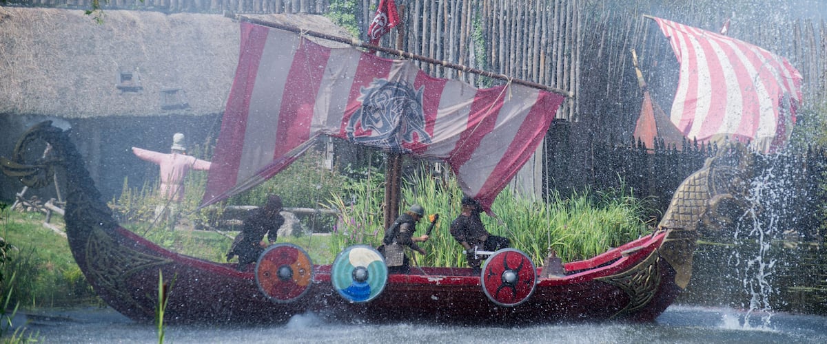 A traditional boat with a red and white striped sail, adorned with a dragon emblem, moving through a water channel.