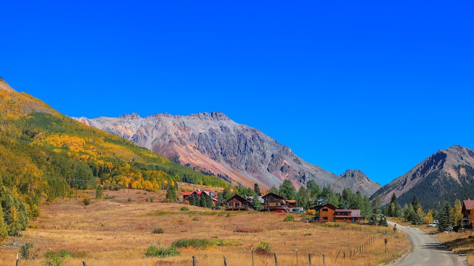A mountain landscape with autumn-colored trees, a clear blue sky, and a road leading through a valley.
