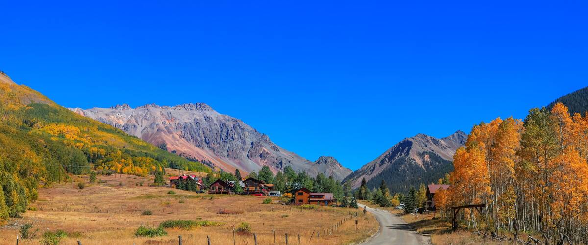 A mountain landscape with autumn-colored trees, a clear blue sky, and a road leading through a valley.