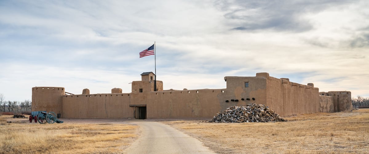 A historic fort with a flag, a dirt road, and dry grassland.