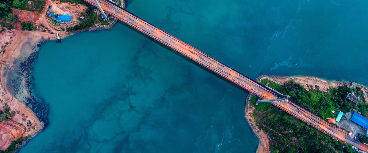 An aerial view of a bridge spanning a body of water, surrounded by greenery and construction sites.