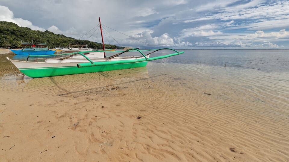 A beach with several boats, clear water, and a cloudy sky.