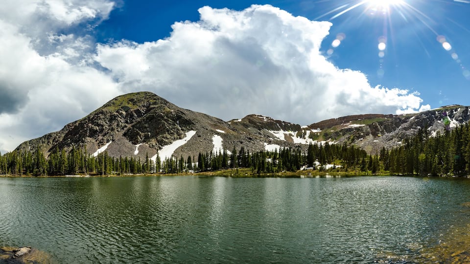 A mountain lake surrounded by dense forests and snow-capped peaks under a partly cloudy sky.