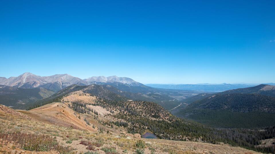 A mountainous landscape with a clear blue sky, a small lake, and dense forests.