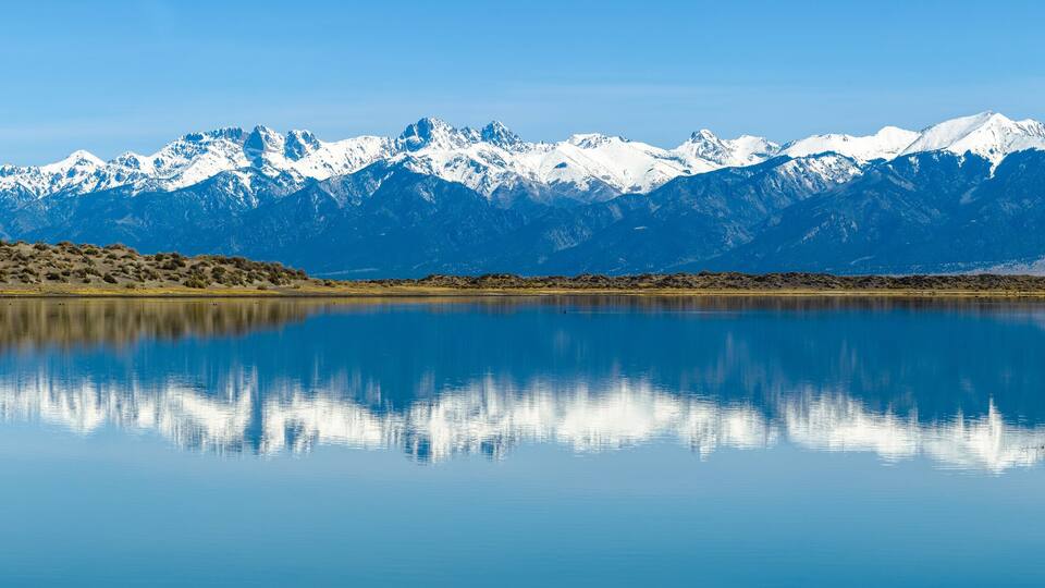 A calm lake reflecting snow-capped mountains under a clear sky.