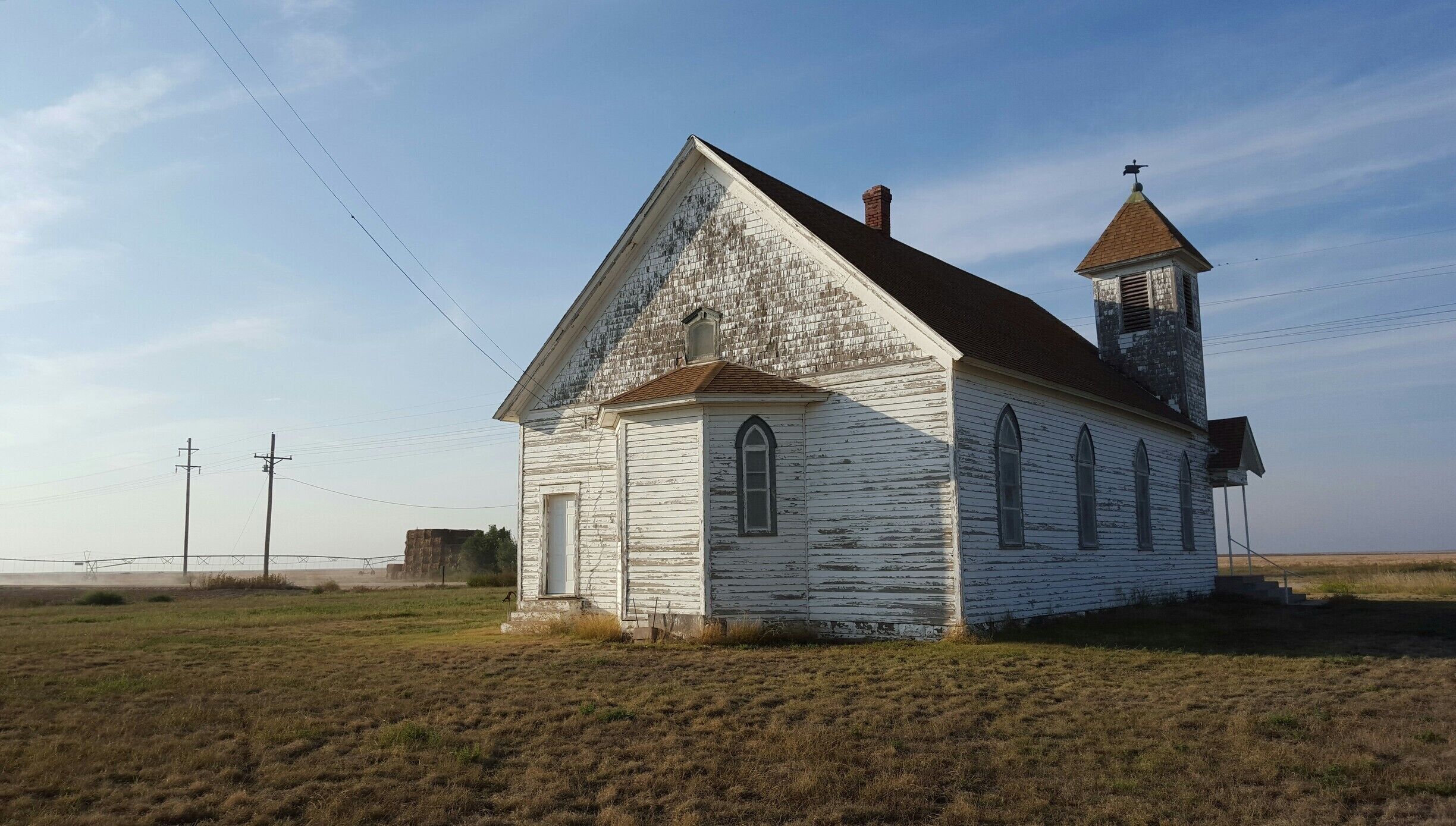 A white church with a steeple and a bell tower, surrounded by open fields and power lines.