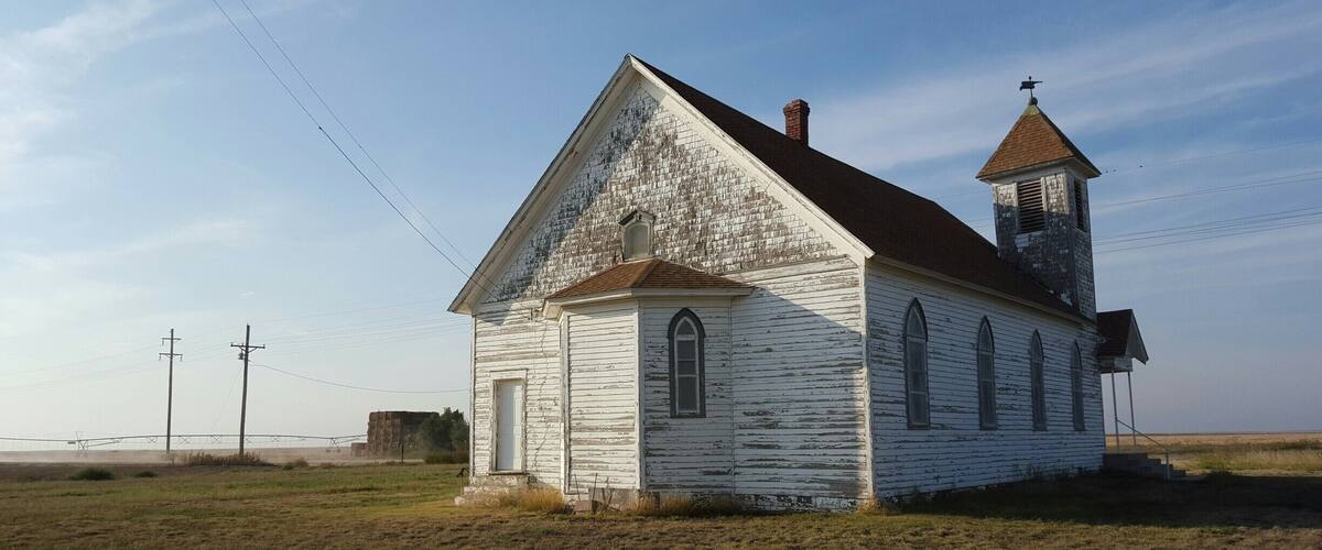 A white church with a steeple and a bell tower, surrounded by open fields and power lines.