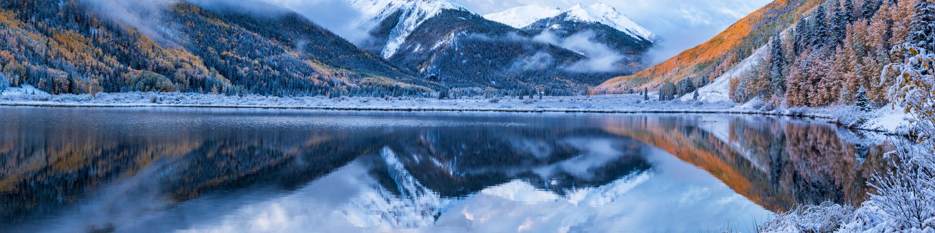 Um lago de montanha tranquilo com árvores cobertas de neve e montanhas ao fundo.