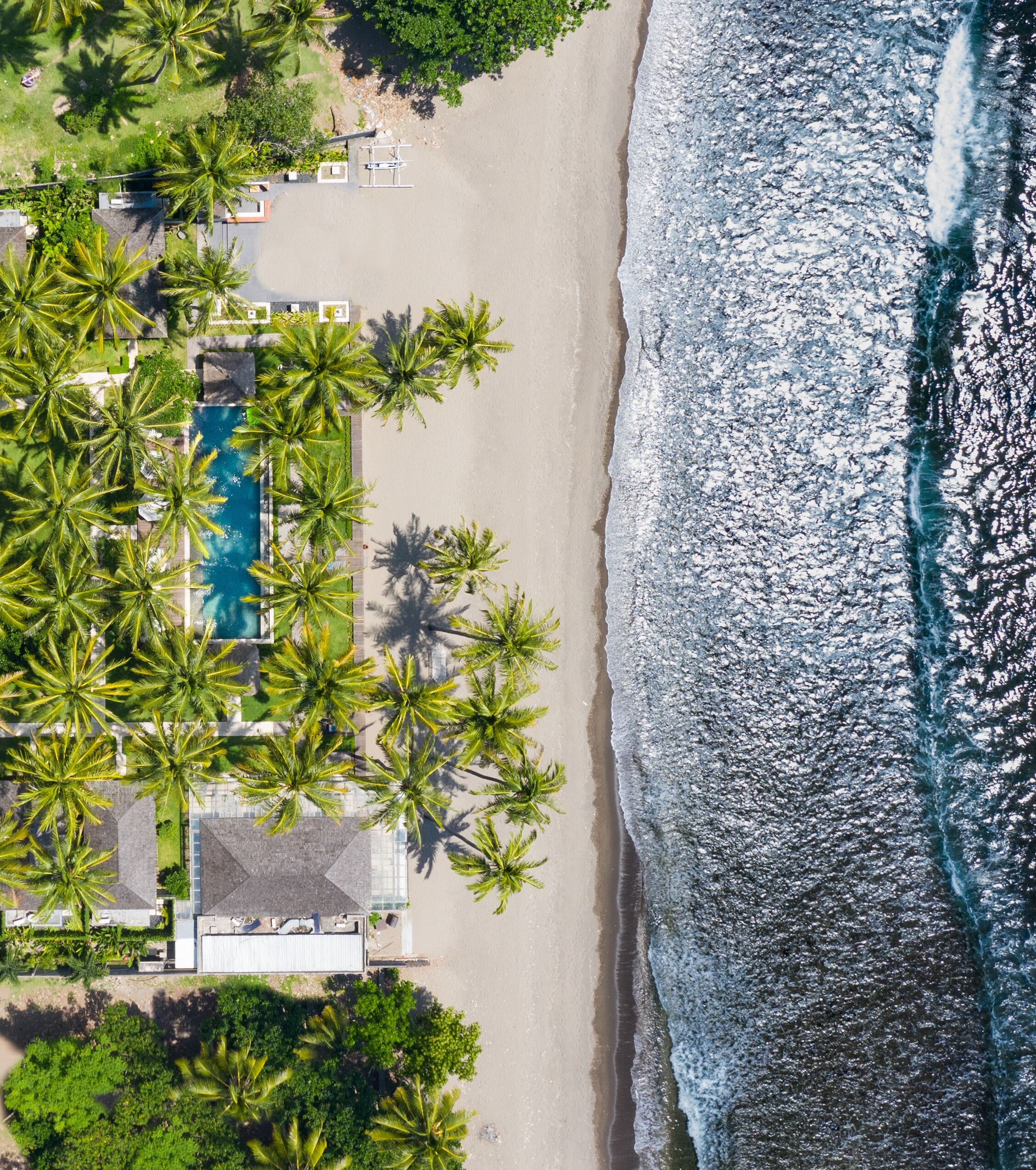 An aerial view of a beach with palm trees, a swimming pool, and a small boat.