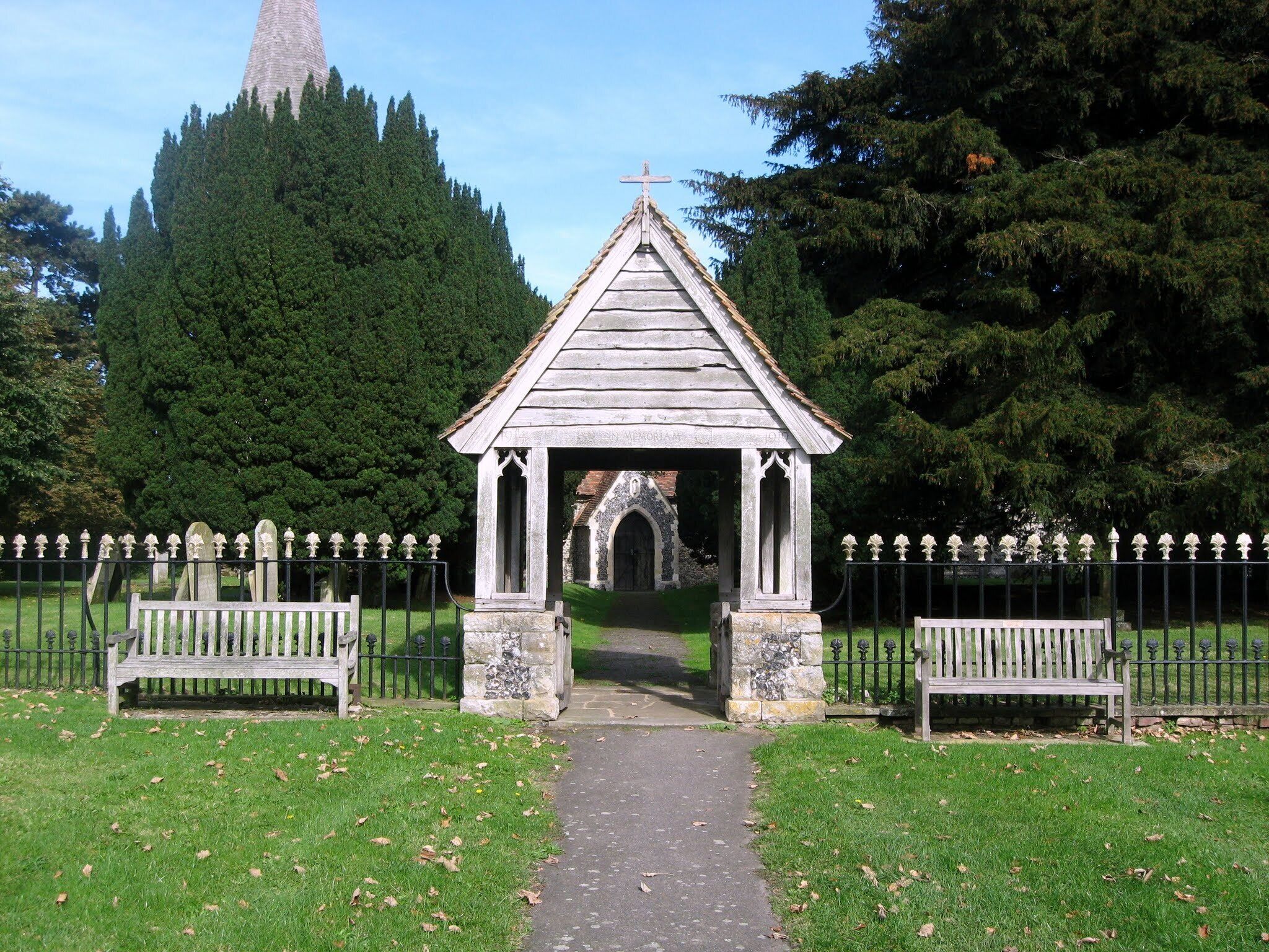 A small wooden chapel with a cross on top, surrounded by a fence and benches, set in a grassy area with trees in the background.