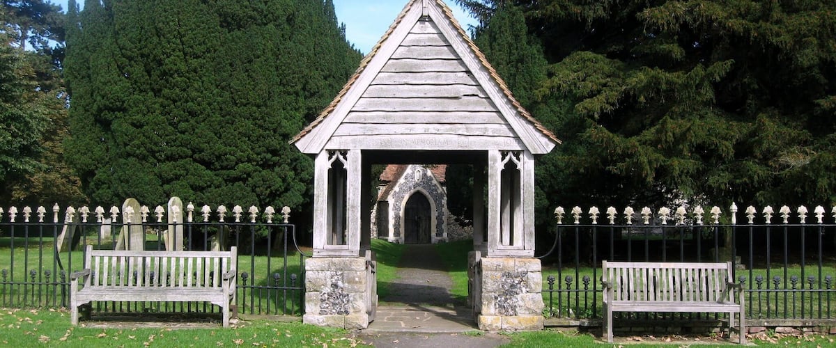 A small wooden chapel with a cross on top, surrounded by a fence and benches, set in a grassy area with trees in the background.
