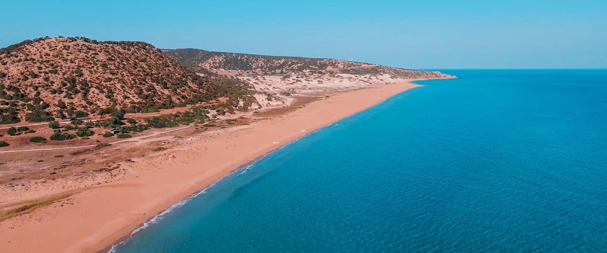 A coastal landscape with a sandy beach, a clear blue sea, and a hilly terrain with sparse vegetation.
