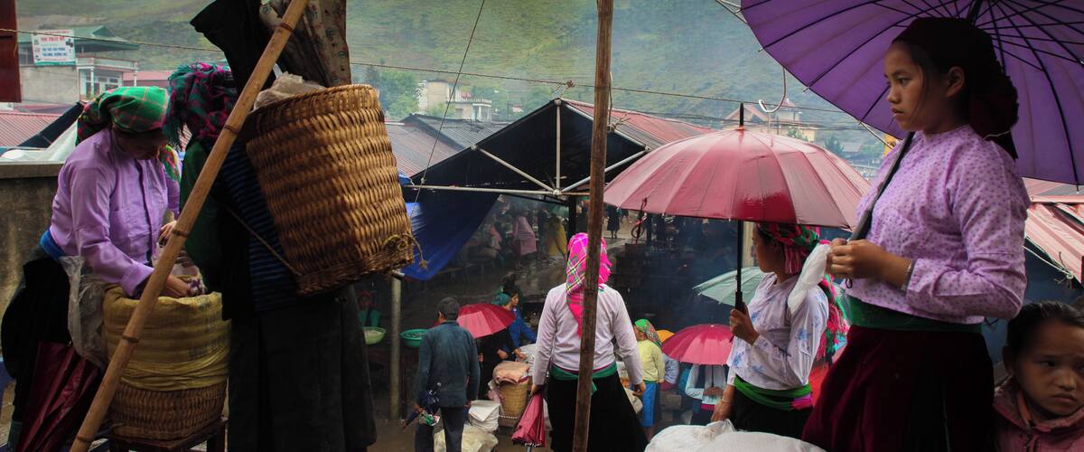 A market scene with people under umbrellas, a woman carrying a basket, and a mountainous background.