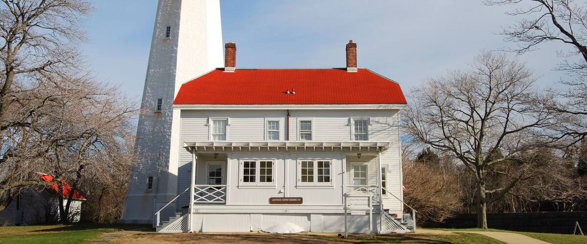 A white lighthouse with a red roof and a white building with a red roof in front of it.