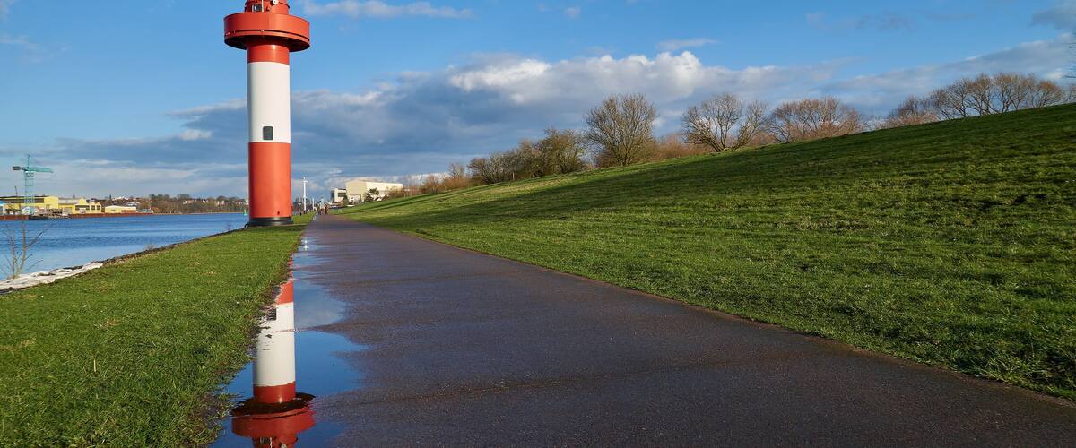 A red and white lighthouse on a wet path beside a grassy hill and a body of water.