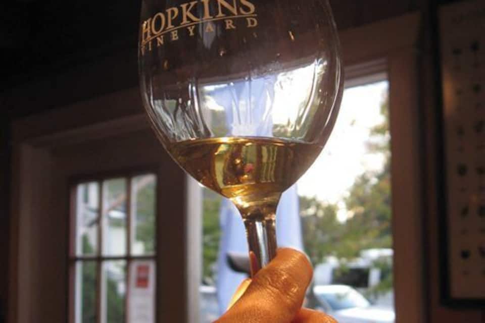 A hand holding a wine glass with the label "Hopkins Vineyard" in a dimly lit room with windows and a door in the background.