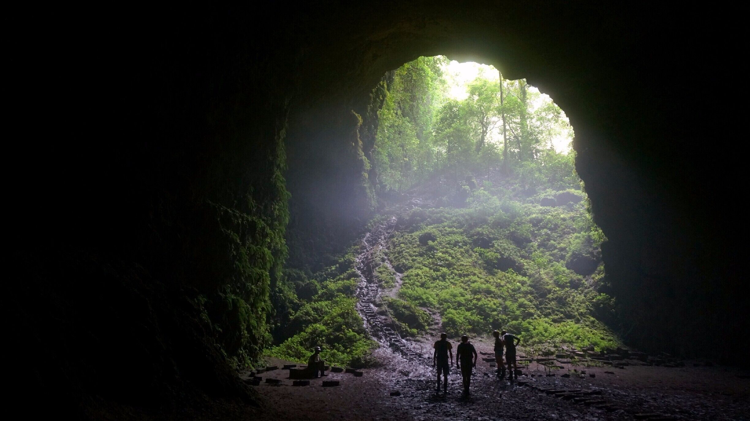 A cave entrance with a waterfall inside, surrounded by lush greenery.