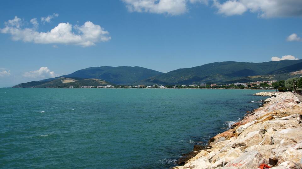 A coastal landscape with a rocky shoreline, calm sea, and distant mountains under a clear sky.