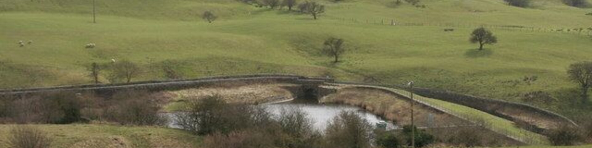Paisagem rural com colinas ondulantes, um pequeno riacho e uma ponte de pedra.