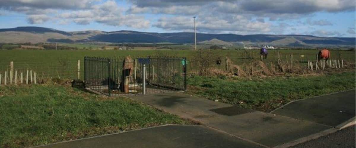 A rural landscape with a fenced area, a road, and distant hills under a partly cloudy sky.