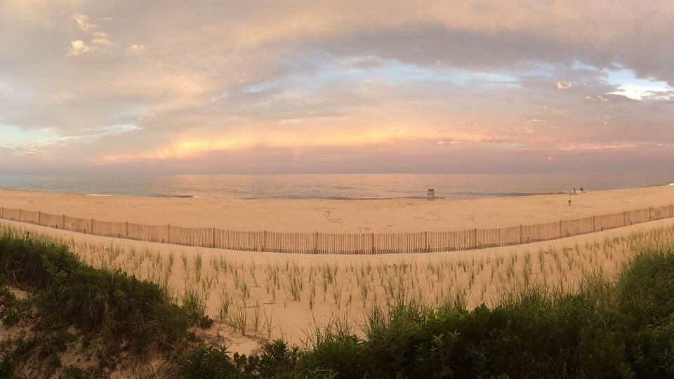 A beach with a wooden fence, grassy dunes, and a cloudy sky.