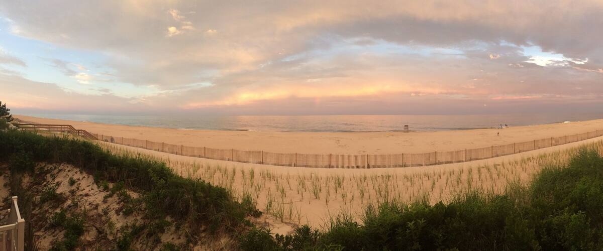 A beach with a wooden fence, grassy dunes, and a cloudy sky.