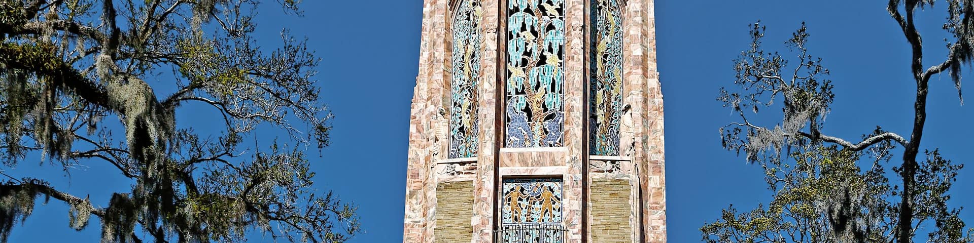 A tall tower with intricate stone carvings and stained glass windows, surrounded by trees and a clear blue sky.