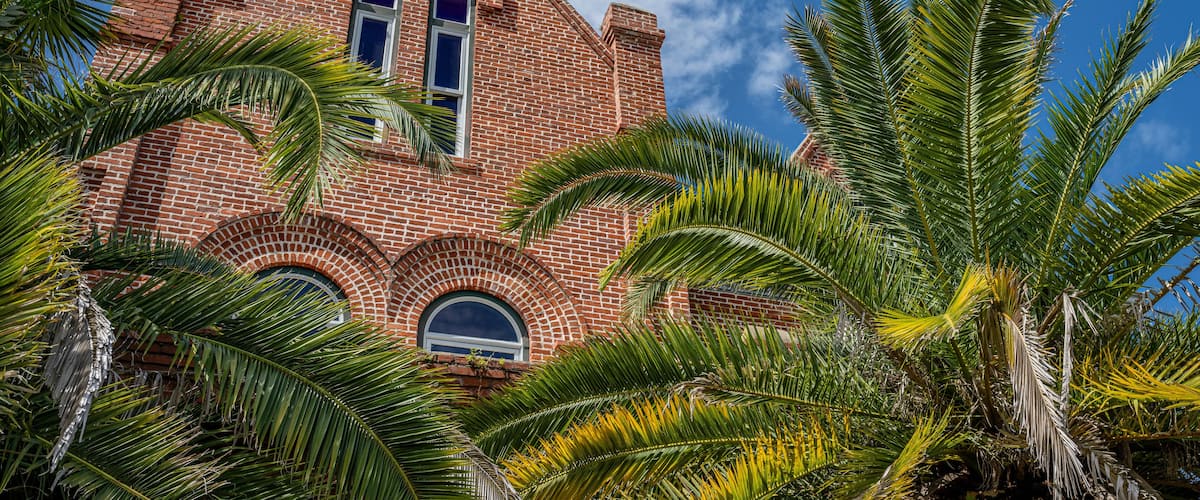 A red brick building with arched windows and a triangular roof, surrounded by palm trees.
