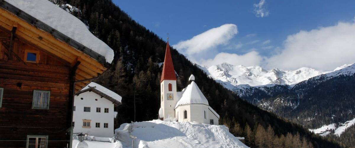 A wooden house, a white church with a red roof, and snow-covered mountains under a clear blue sky.