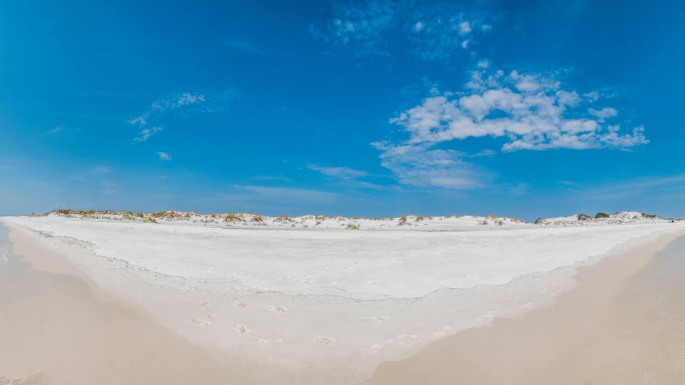 A wide sandy beach with clear shallow waters and dunes in the background.